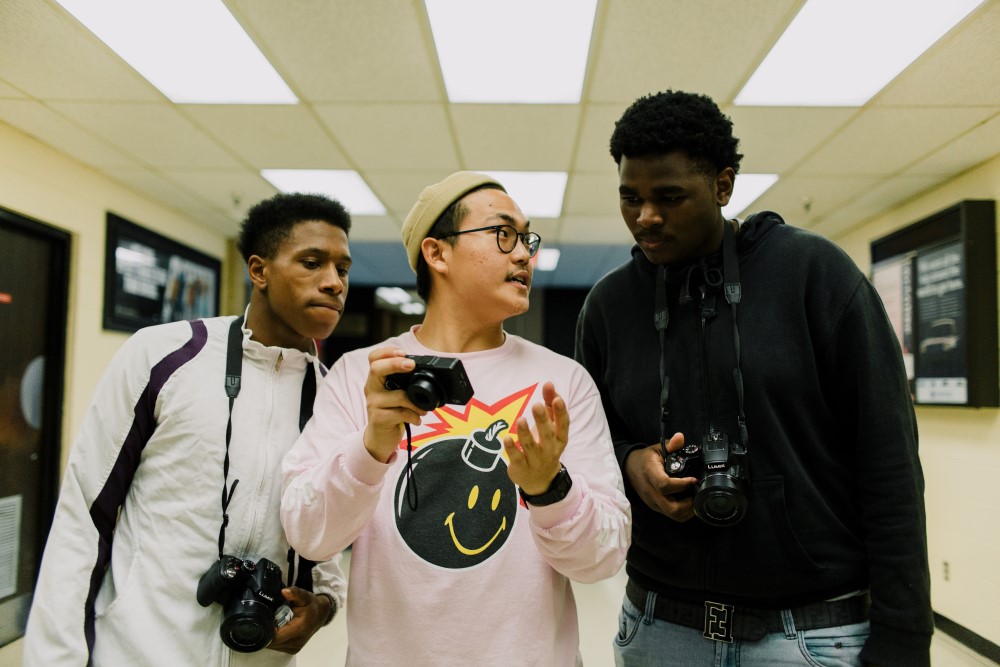 Two students holding cameras look over Joey Park’s shoulder as he explains his camera screen to them.