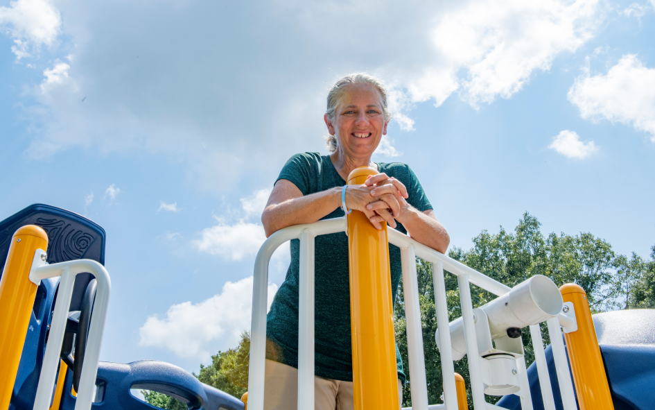marybeth smiles looking downward from the playground equipment with a partly cloudy sky as her background.