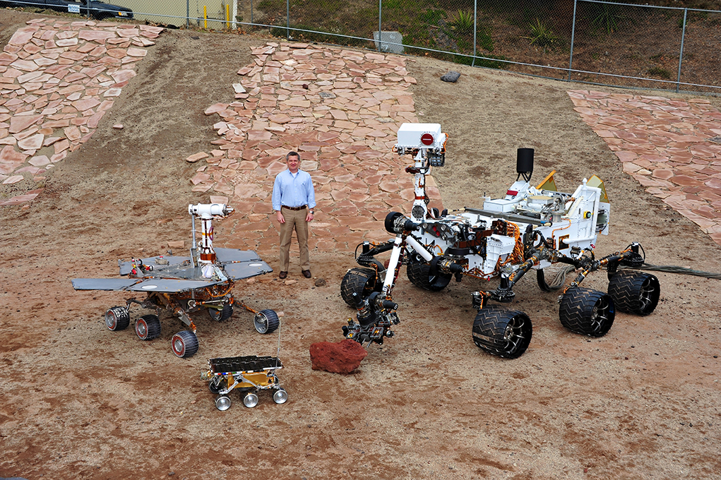 Keith Comeaux standing in an open field with several Mars rovers