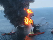 an oil well on fire with boats surrounding it and trying to put the fire out with hoses