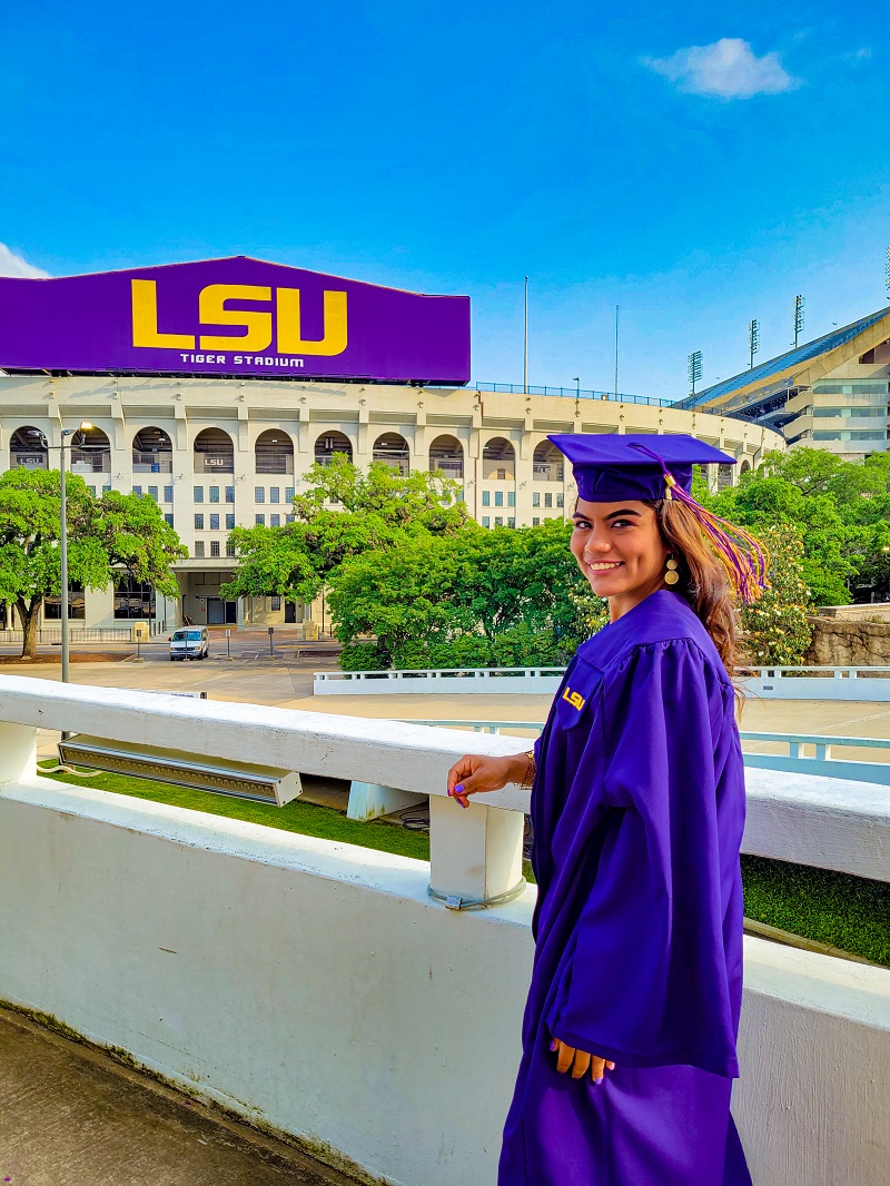 a woman in a graduation cap and gown smiles in front of Tiger Stadium