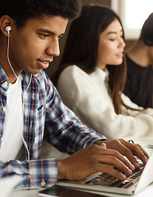 Photo of a young man working on a laptop with earphones in his ears. A young woman is sitting next to him.
