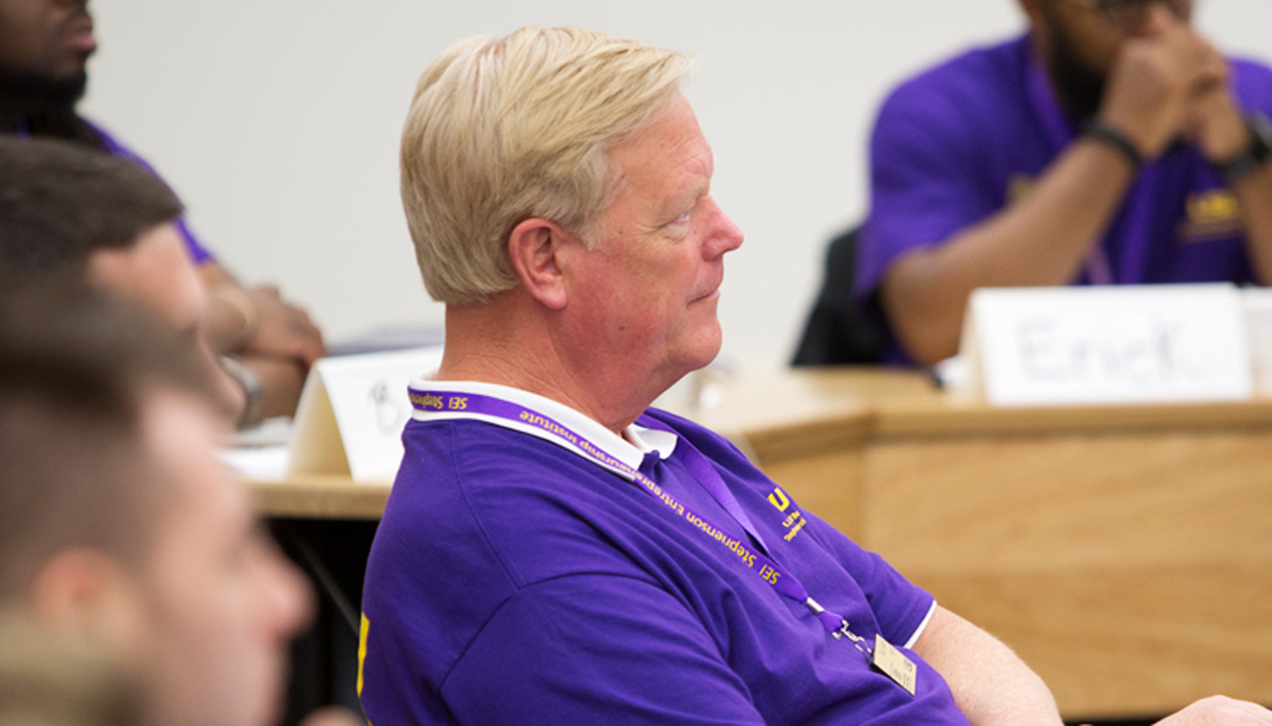 Man in purple shirt listens in classroom with arms crossed. 