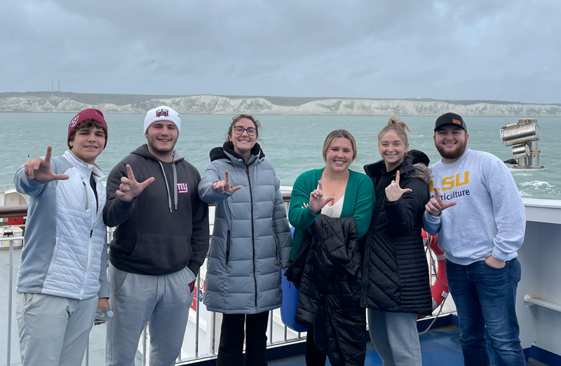 A small group of students stand atop a ferry crossing the English Channel. The White Cliffs of Dover are in the background. It looks to be a cold day. Students hold their hands in an L shape to show their LSU spirit.