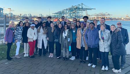 A large group of students including many LSU Tigers pose for a group portrait at the Port of Rotterdam in the Netherlands. It appears to be a cold day as many wear jackets. All are smiling and looking at the camera. A large crane appears in the background. 