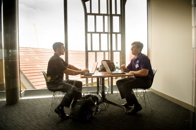 Two male students talk at a table with their laptops.